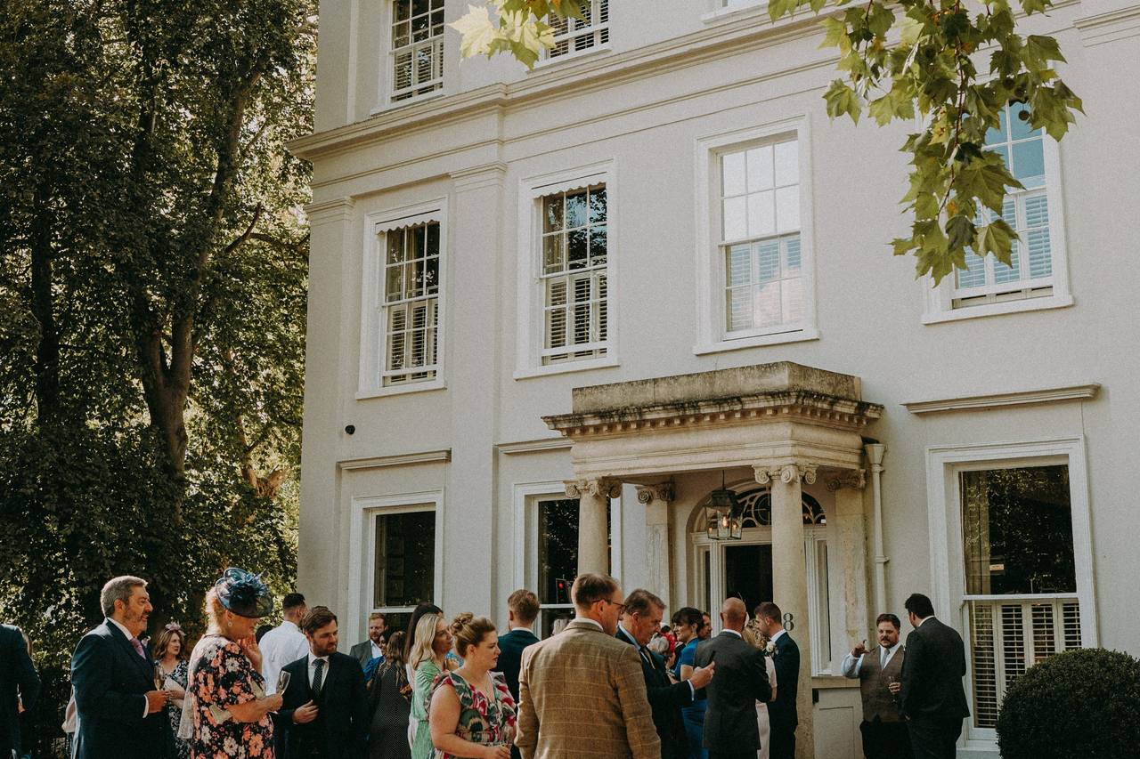 A joyful wedding party gathers in front of a large white house, showcasing elegant outfits and a festive atmosphere.