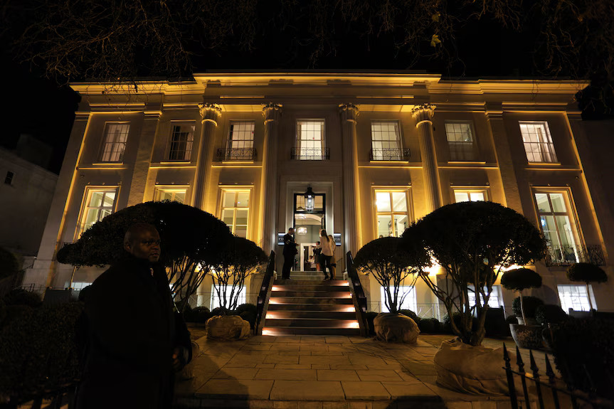 A man stands in front of a large mansion illuminated at night, showcasing its architectural details against the dark sky.